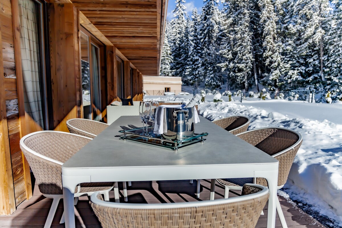 A wooden cabin with a dining table and wicker chairs, surrounded by snow-covered trees.