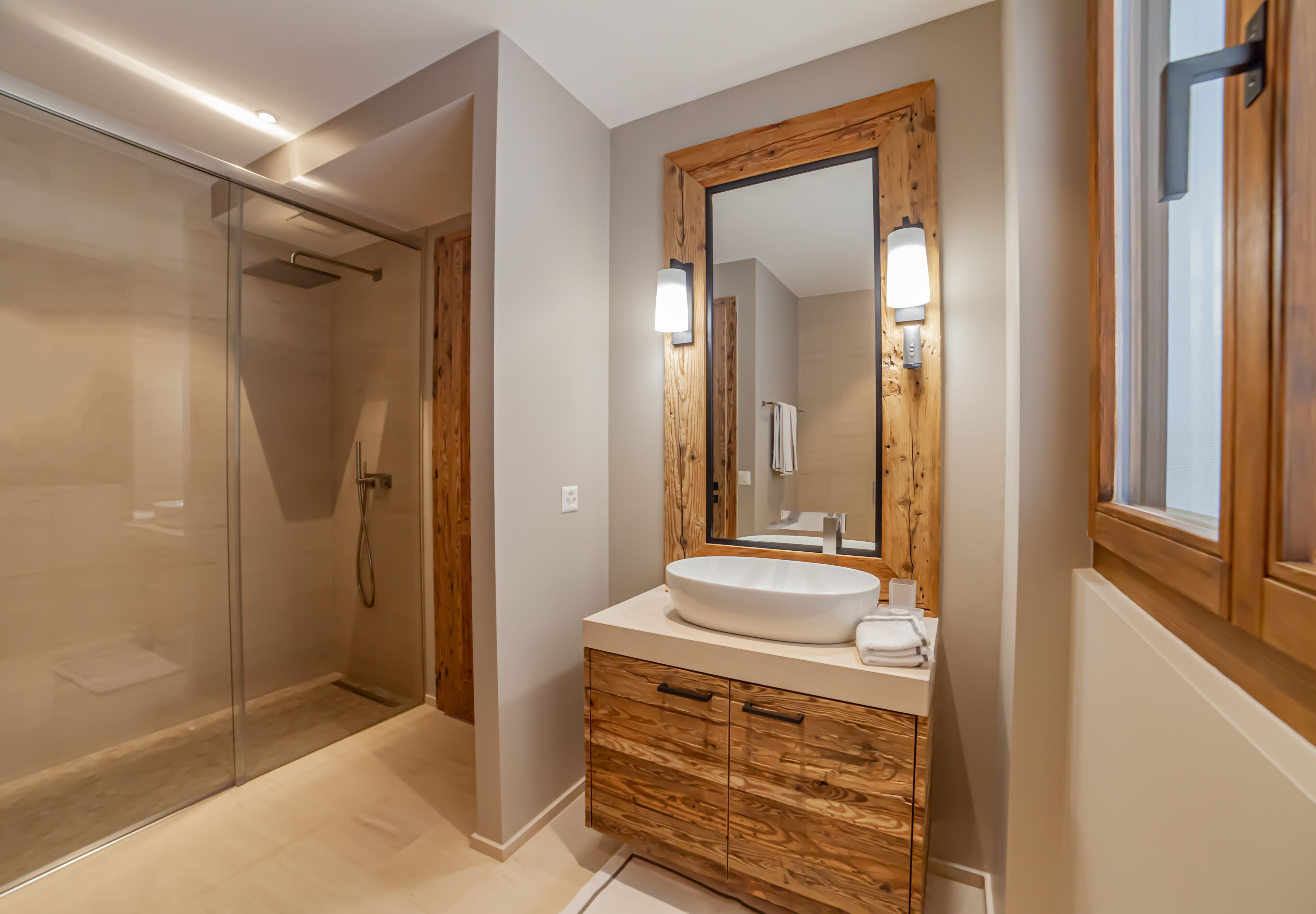Ein modernes Badezimmer mit Duschkabine aus Glas, einem Holzmöbelkorpus und einem großen Spiegel sowie einem weißen Waschbecken.