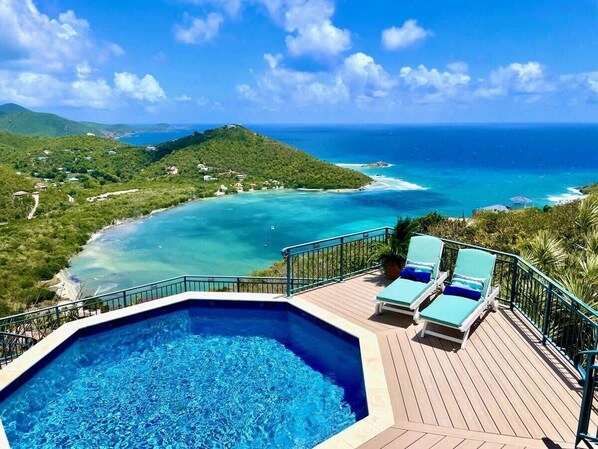 A swimming pool with lounge chairs, a deck, and a view of a coastal area with clear blue water and green hills.