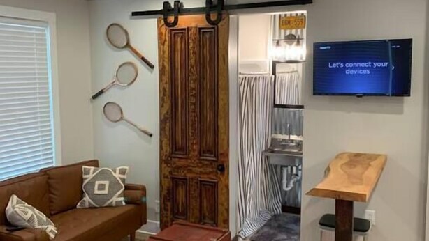 A living room with a brown sofa, a red ottoman, a television mounted on the wall, and a wooden stand. - Refined Studio, New Construction, 1 Mile From Town (Pittsburgh)