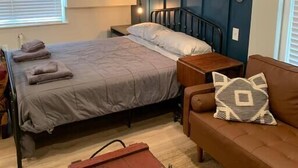 A bedroom with a bed, a nightstand, a dresser, a window with blinds, and a wall-mounted air conditioner. - Refined Studio, New Construction, 1 Mile From Town (Pittsburgh)