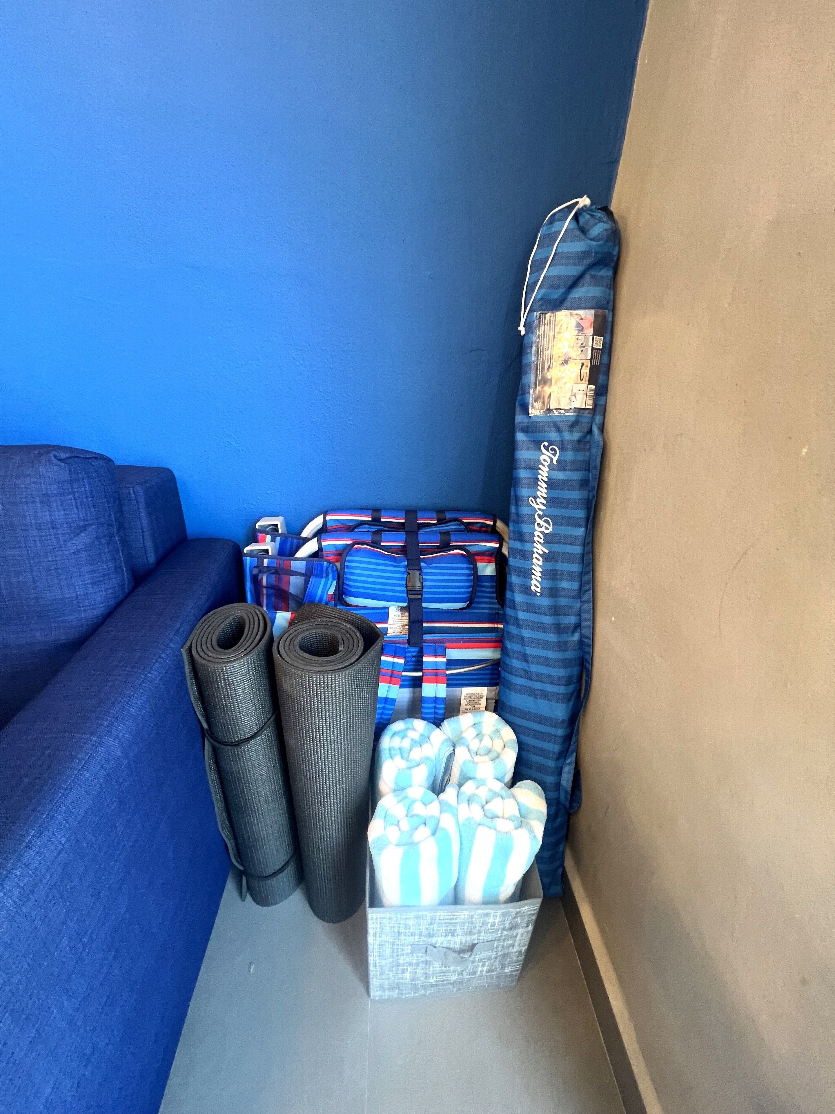Un coin de pièce avec un mur bleu, un canapé gris et une boîte de rangement contenant des tapis enroulés et une couverture à rayures bleues et blanches.