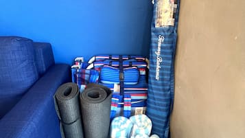 Een hoek van een kamer met een blauwe muur, een grijze bank en een opbergkist met opgerolde matten en een blauw-witte strepen deken.