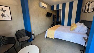 A hotel room with a bed, a chair, a small table, an air conditioning unit, and a ceiling light fixture.