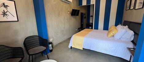 A hotel room with a bed, a chair, a small table, an air conditioning unit, and a ceiling light fixture.