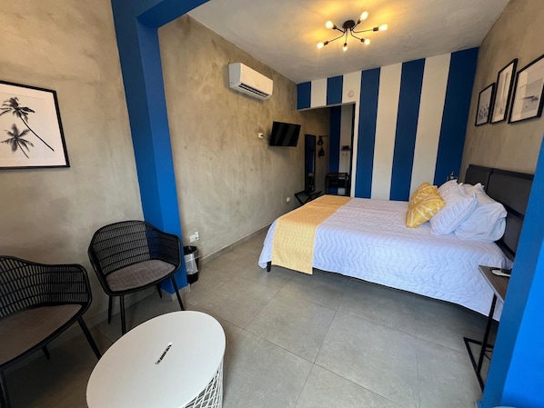 Habitación de hotel con cama, una silla, una mesita pequeña, equipo de aire acondicionado y lámpara en el techo.