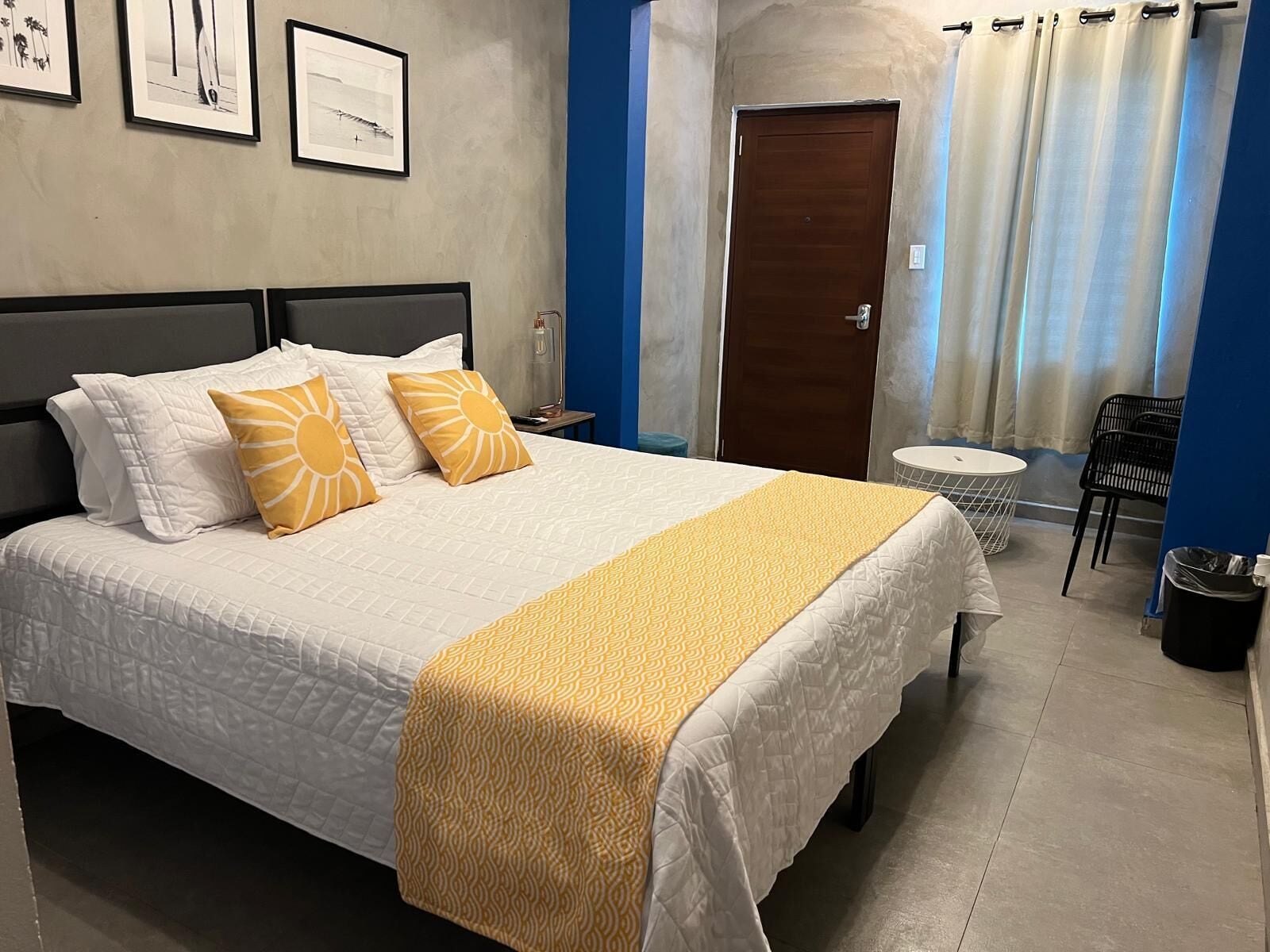 Un lit bien fait, recouvert d’une couvre-lit blanche et jaune, avec une tête de lit en bois et deux oreillers ornés de motifs solaires. On aperçoit une petite table ronde avec une chaise à côté du lit, et une poubelle est placée dans un coin.