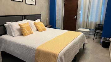 Un lit bien fait, recouvert d’une couvre-lit blanche et jaune, avec une tête de lit en bois et deux oreillers ornés de motifs solaires. On aperçoit une petite table ronde avec une chaise à côté du lit, et une poubelle est placée dans un coin.