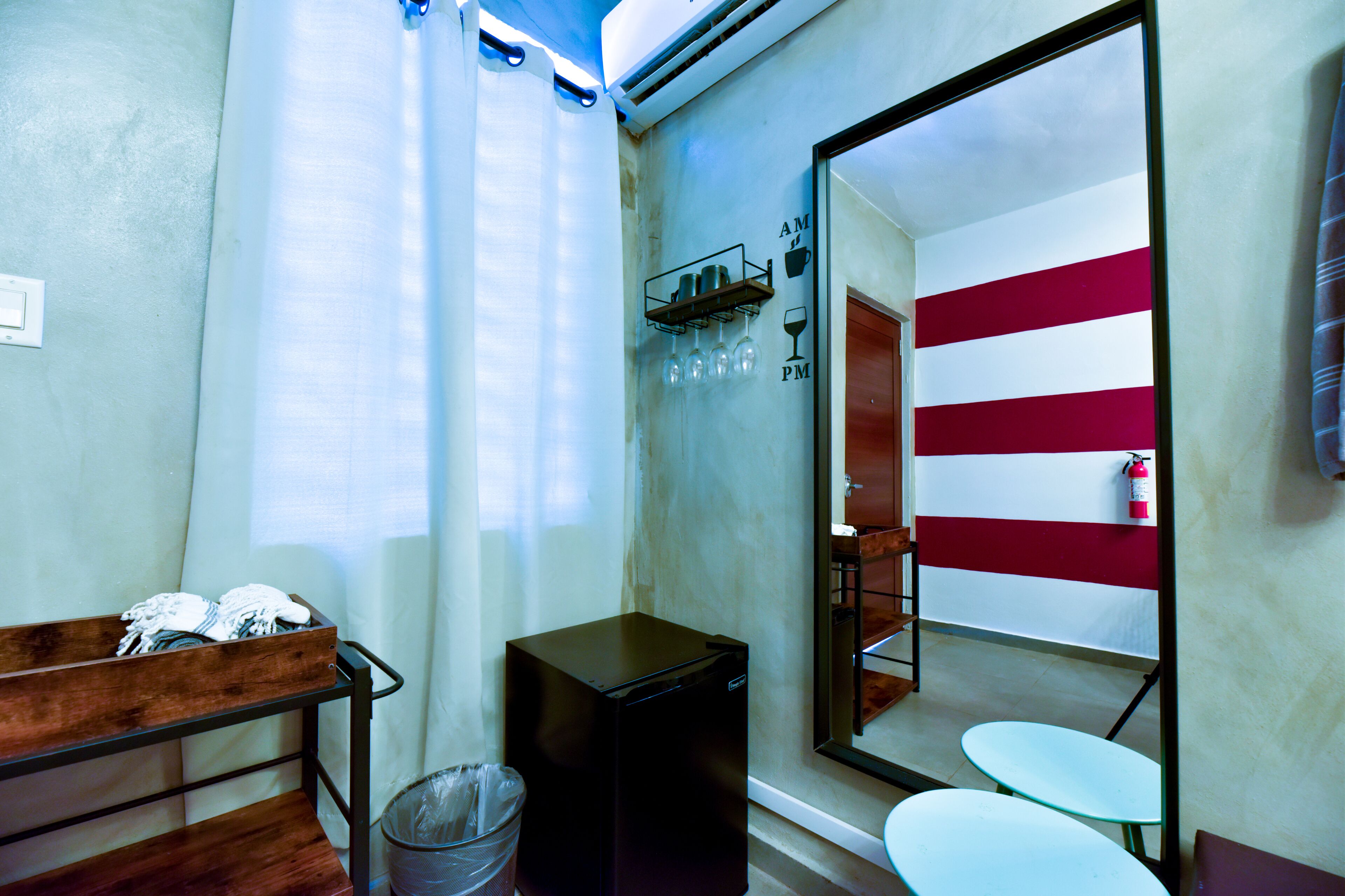 A compact hotel room with a small kitchenette, a wooden bench with towels, a trash bin, and a mirror reflecting a bathroom with a red and white striped wall.