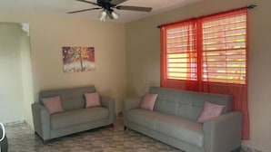 A living room with two grey sofas, a ceiling fan, a painting on the wall, and a window with red curtains. - Private Country house 5 miles from beach (Aguada)