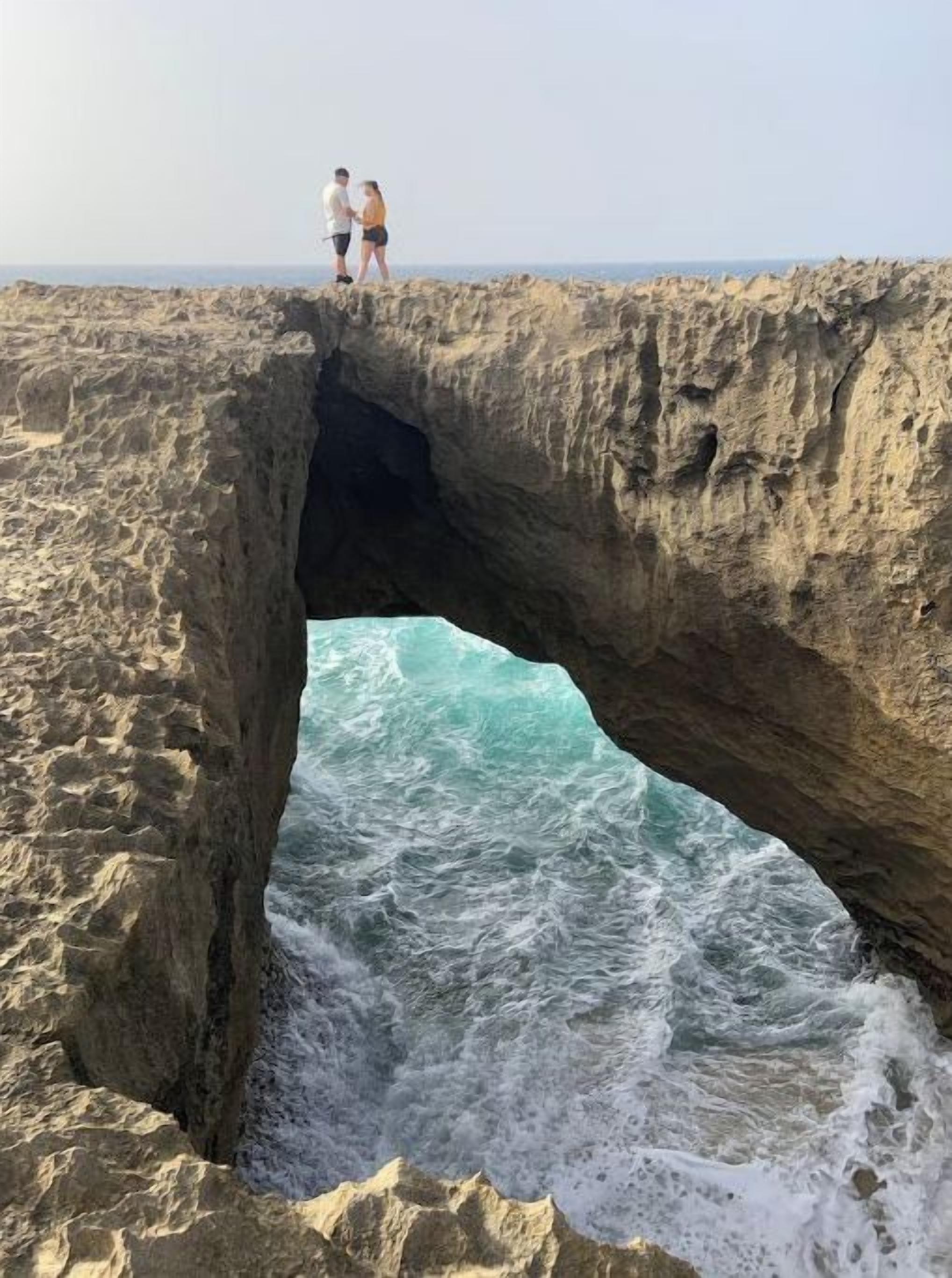 A couple standing on a rocky cliff overlooking a turbulent sea through a natural arch formation.