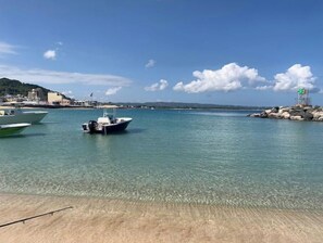 Freie Weitsicht auf das Wasser mit vor Anker liegenden Booten, ein Sandstrand und ein Felsensteg.