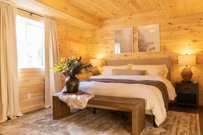 Un dormitorio de cabaña de madera con una cama, lámparas de noche, una ventana con cortinas y un cuadro colgado en la pared.