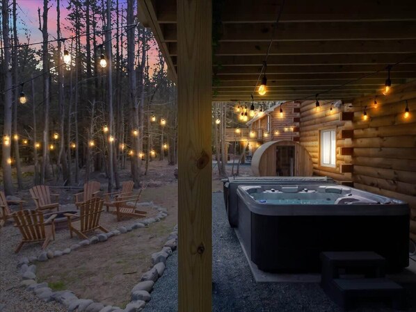Una cabaña de troncos con jacuzzi, mobiliario exterior e iluminación con luces colgantes.