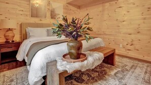 Interior de una cabaña de madera con una cama, un jarrón con flores, una lámpara y una alfombra.