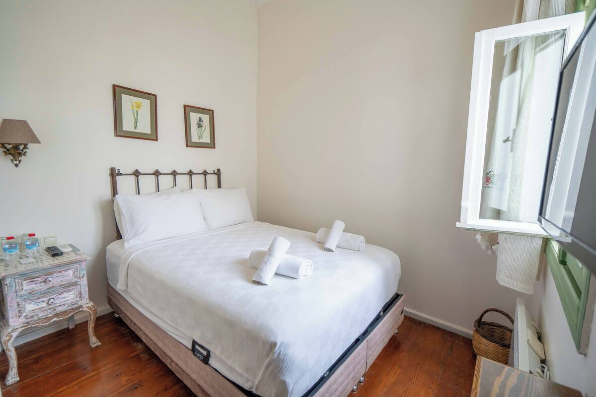 A neatly made bed with white linens and two rolled towels, a small bedside table with a lamp, and two framed pictures on the wall.