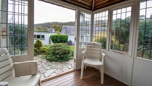A conservatory with stone flooring, white chairs, and a view of a house and greenery.