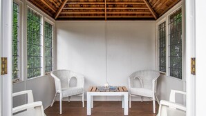 A room with two white wicker chairs, a small wooden table, and large windows with a view of greenery.