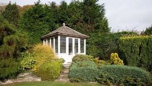 A small white gazebo with a tiled roof, surrounded by lush greenery and various shrubs.