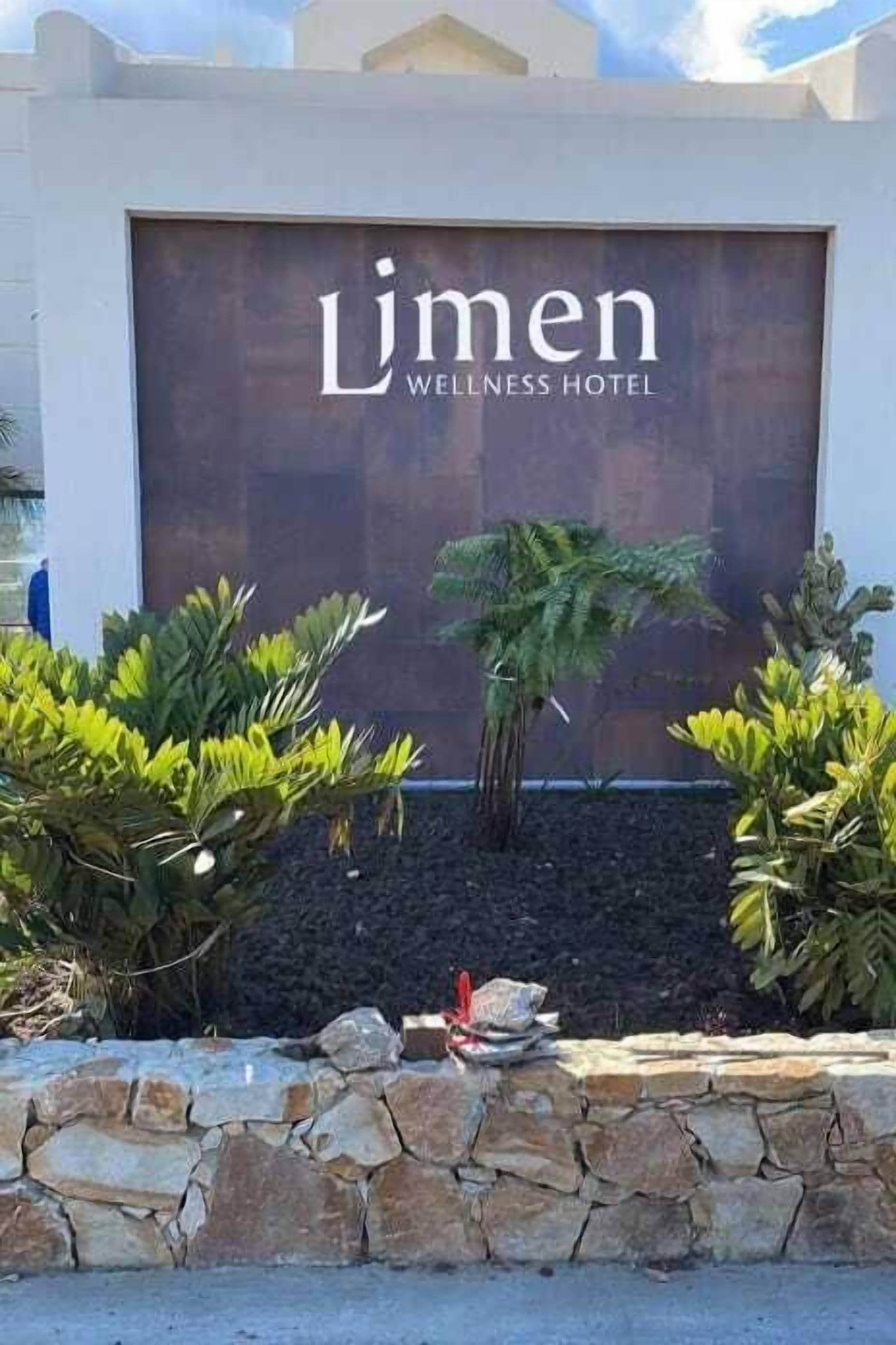 A sign for the Lumen Wellness Hotel with a stone wall and tropical plants in front.