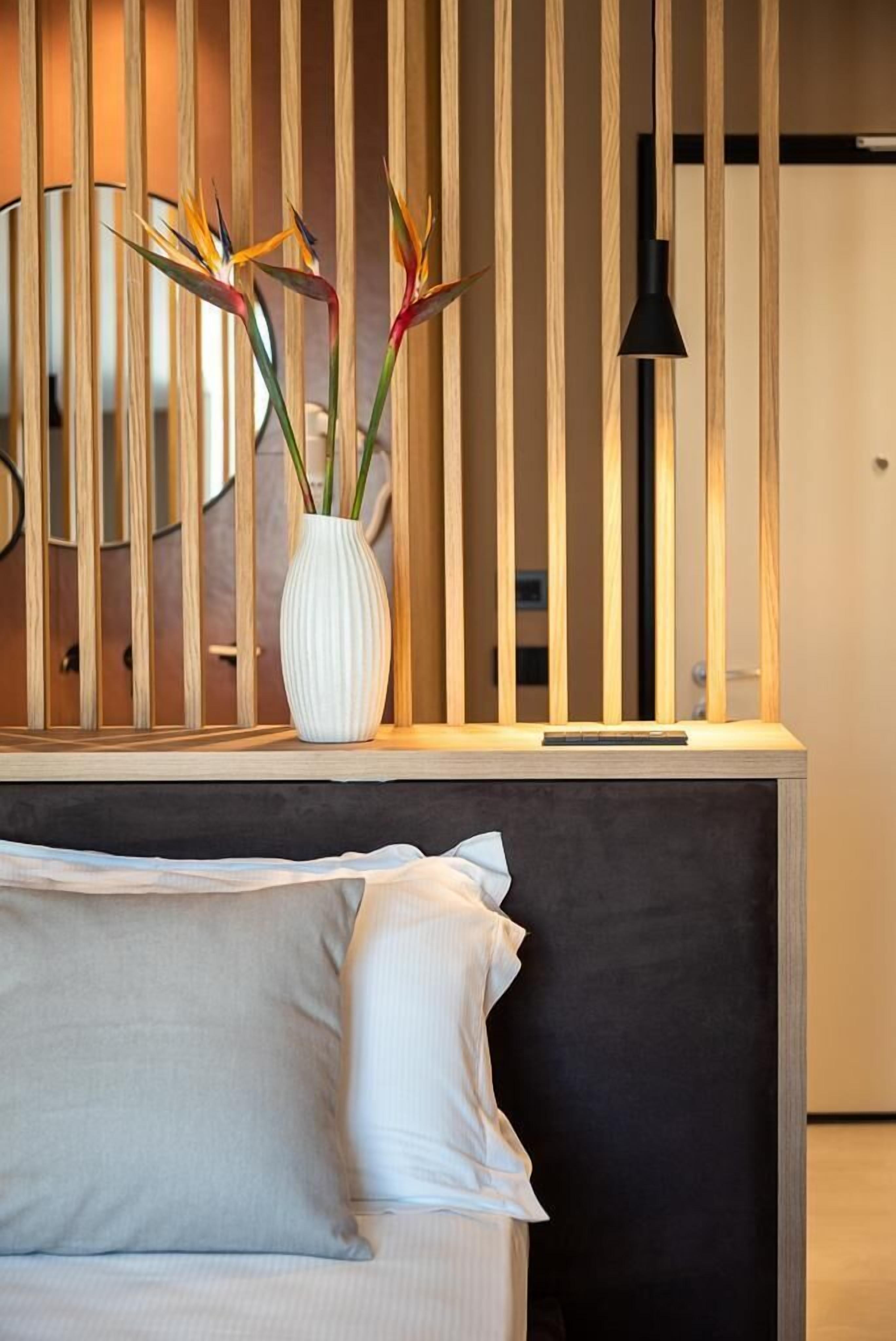 Une chambre moderne avec un paravent en lattes de bois, un vase blanc orné de fleurs, une lampe de sol noire et un lit avec une literie blanche.