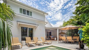 A pool area with lounge chairs and a dining table, surrounded by a white house and lush greenery.