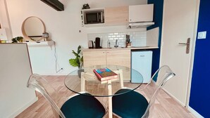 Une cuisine compacte équipée d’un four à micro-ondes, d’une machine à café et d’un petit réfrigérateur. On y trouve une table à manger avec deux chaises, et un livre est posé dessus.