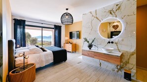 Un dormitorio moderno con cama, mesita de noche, televisor, baño con pared de mármol, lavamanos y espejo.