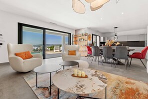 Una sala de estar moderna con un ventanal grande, un comedor con una silla roja y una cocina al fondo.