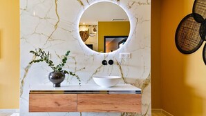 Un baño con pared de mármol, un espejo redondo, un lavamanos blanco, un mueble de madera y una planta.
