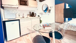 A modern kitchen with white cabinets, a microwave, and a sink. There is a glass table with chairs and a book. A small refrigerator is visible in the corner.