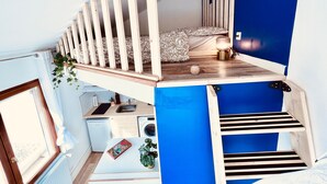 A loft bedroom with a sloped ceiling, a built-in bed, a small kitchen area, and a blue accent wall. - De la Terre à la Lune (Amiens)
