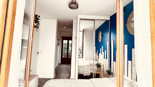 A bedroom with a large bed, a small table with a plant, and a mirror. There is a door leading to another room and a view of the outside. - L'Ile Mystérieuse - Studio avec Terrasse (Amiens)