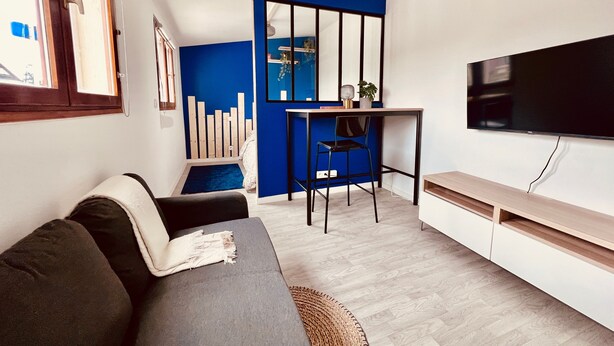 A modern living room with a grey sofa, a small table, a TV, and a blue accent wall. - Tour du monde en 80 jours - Studio avec Terrasse (Amiens)