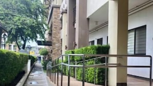 Un paseo techado con escalones que conducen a la entrada de un edificio, bordeado por vegetación y una barandilla metálica.