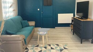 Una sala de estar con un sofá color turquesa, una mesita redonda pequeña, un televisor sobre un mueble de madera y una puerta con un tablón de anuncios.