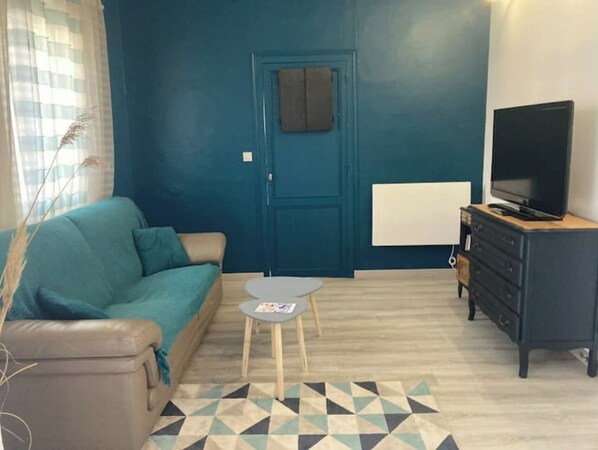 Una sala de estar con un sofá color turquesa, una mesita redonda pequeña, un televisor sobre un mueble de madera y una puerta con un tablón de anuncios.