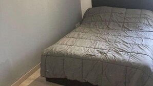 Una cama pequeña y bien hecha con una colcha gris en una habitación con paredes grises claras y suelo de madera.