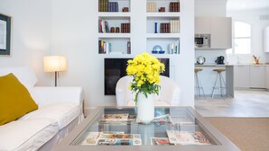 A modern living room with a glass coffee table displaying magazines, a white sofa with a yellow pillow, a white armchair, a bookshelf, and a kitchen area in the background. - Adriano 2.1 by Home at Homes (Sevilla)