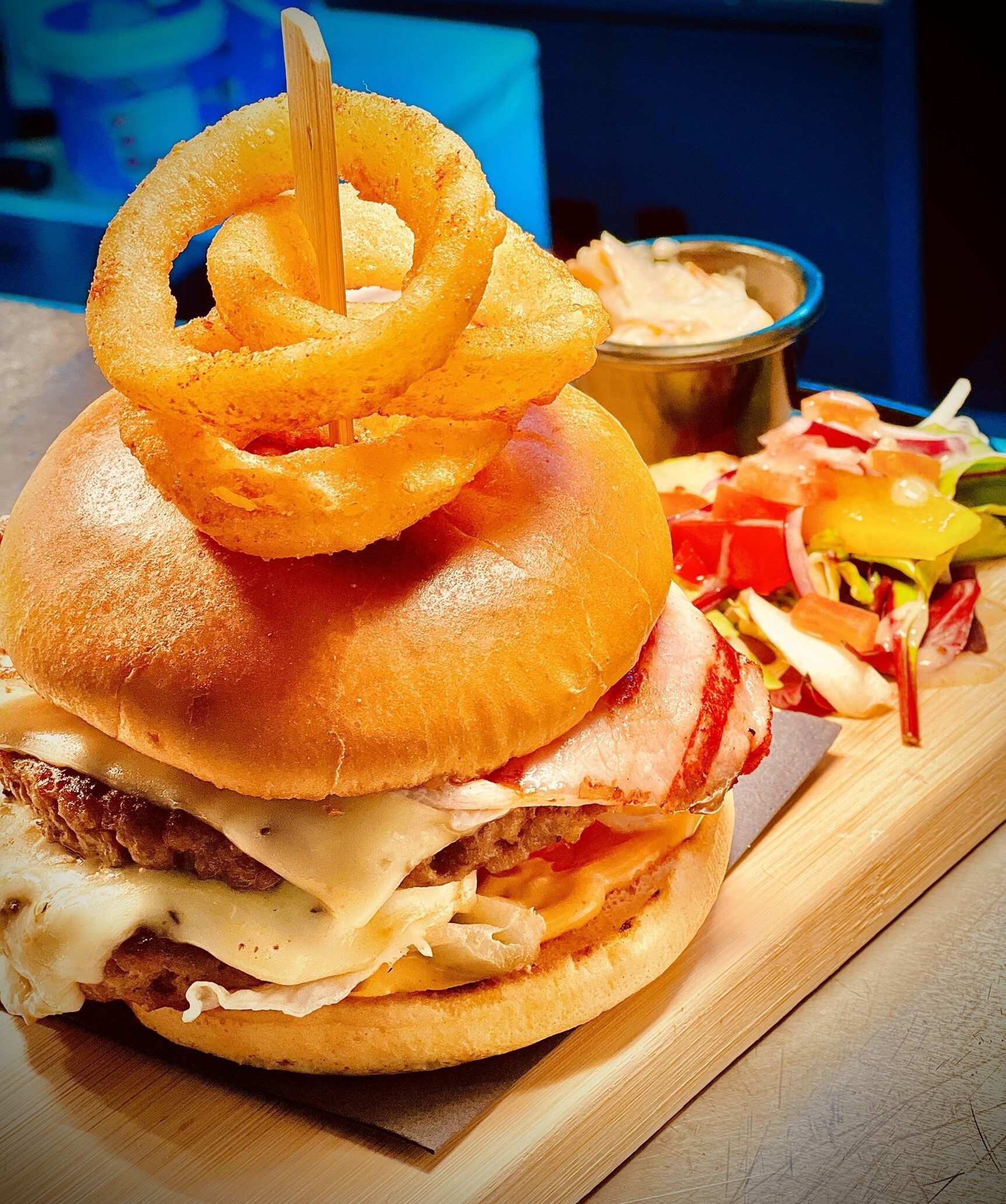 A burger with onion rings on top, a side salad, and a small container of sauce on a wooden board.