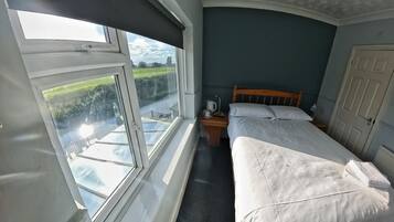 Ein Zimmer mit einem Bett, einem Nachttisch, einem Fenster mit Blick auf eine grüne Landschaft und einer Tür.