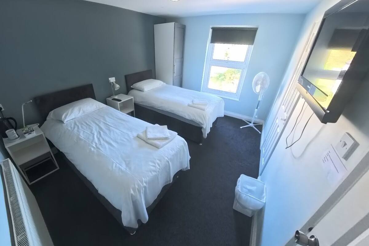 A hotel room with two single beds, a desk with a chair, a standing fan, a window with blinds, and a TV mounted on the wall.