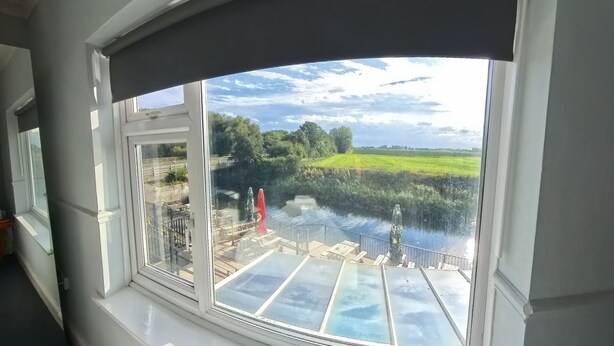 Blick aus einem Zimmer mit großem Fenster auf einen Fluss, eine Wiese und eine Brücke.