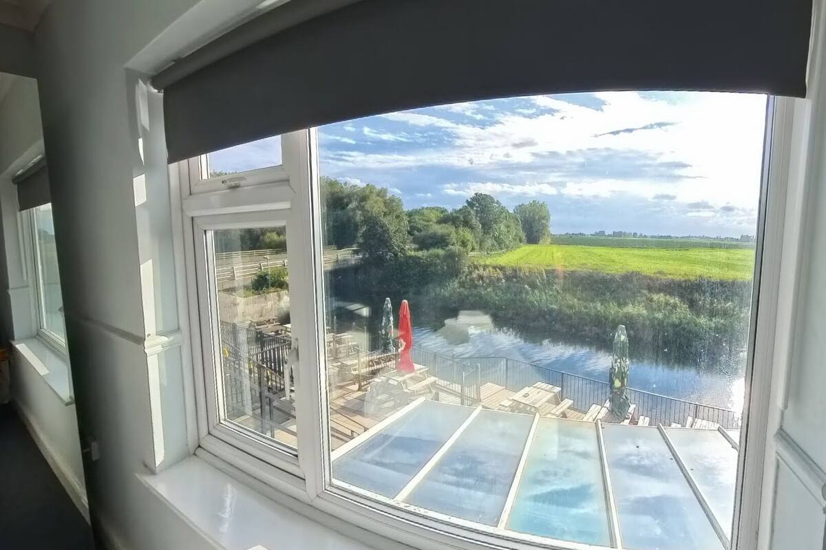 A view from a room with a large window overlooking a river, a grassy field, and a bridge.