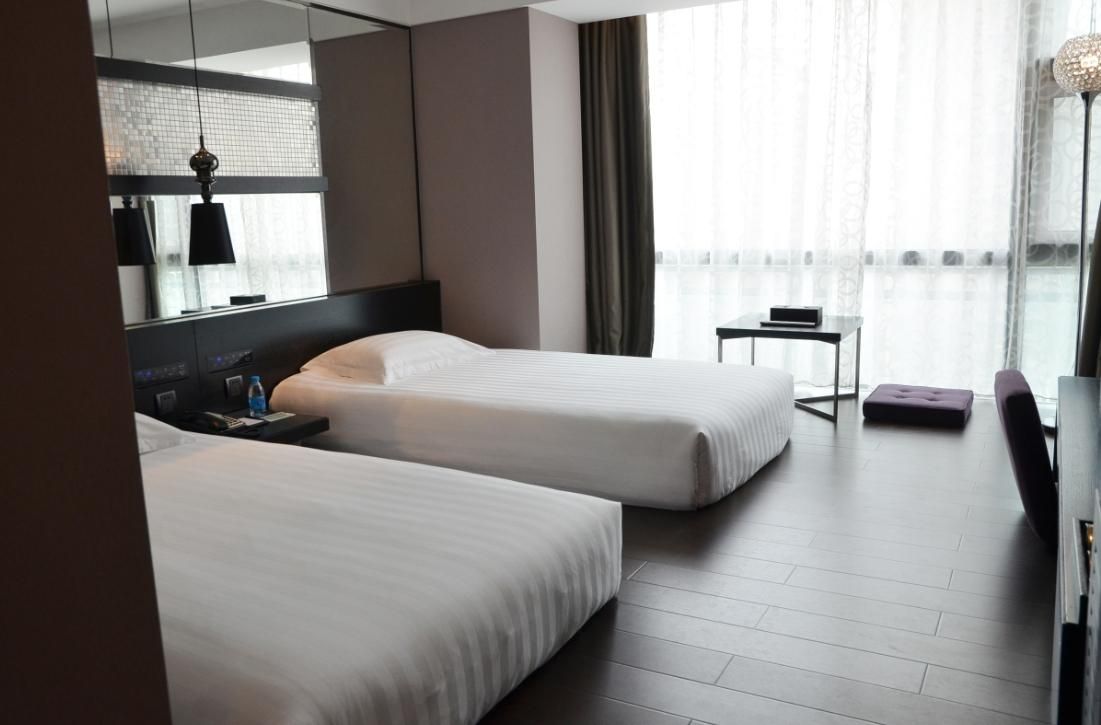 A hotel room with two beds, a small table, a chair, and a large window with curtains.