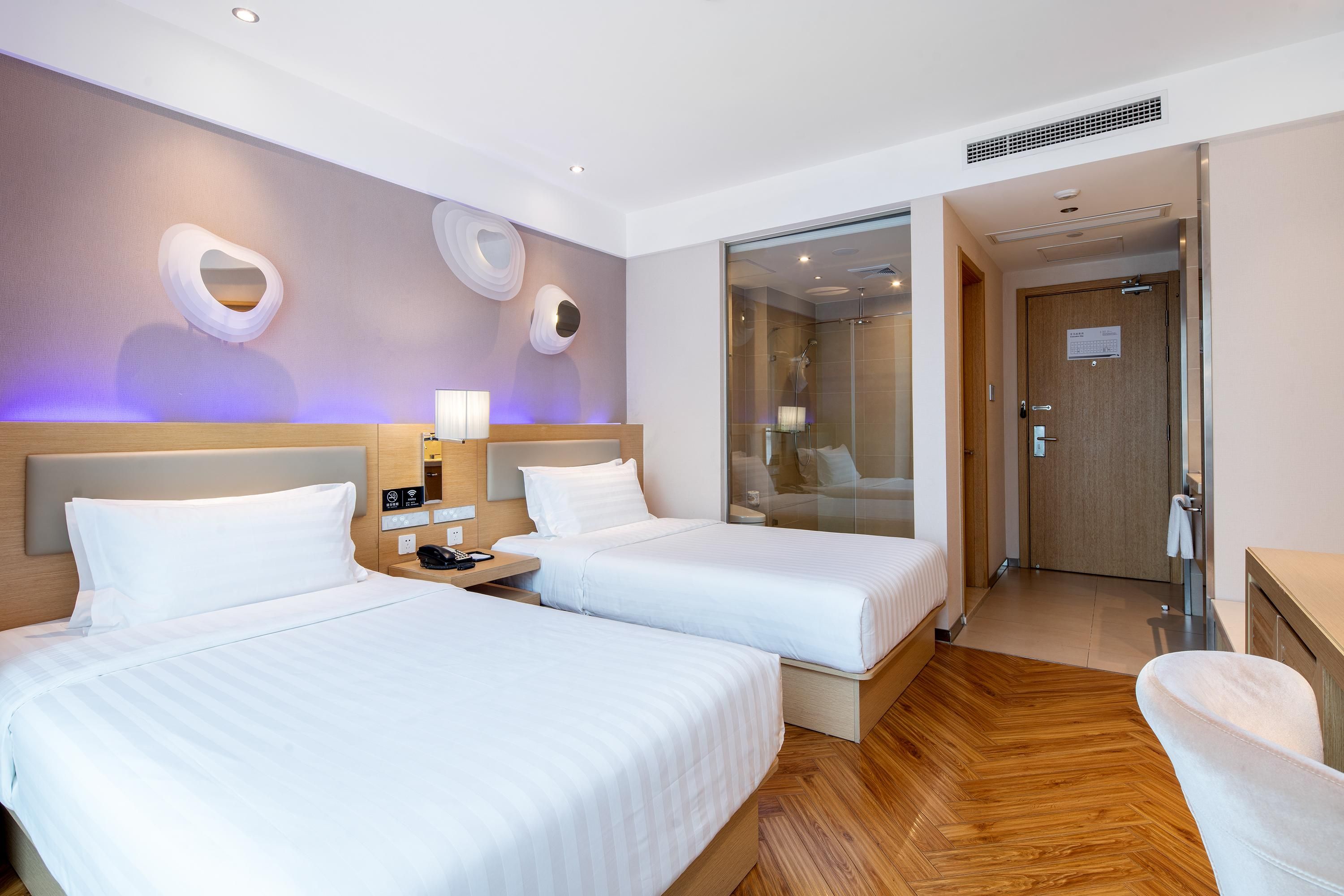 A hotel room with two beds, a wooden floor, a bathroom with a glass shower, and a desk with a chair.