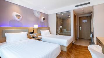 A hotel room with two beds, a wooden floor, a bathroom with a glass shower, and a desk with a chair.