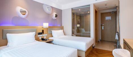 A hotel room with two beds, a wooden floor, a bathroom with a glass shower, and a desk with a chair.