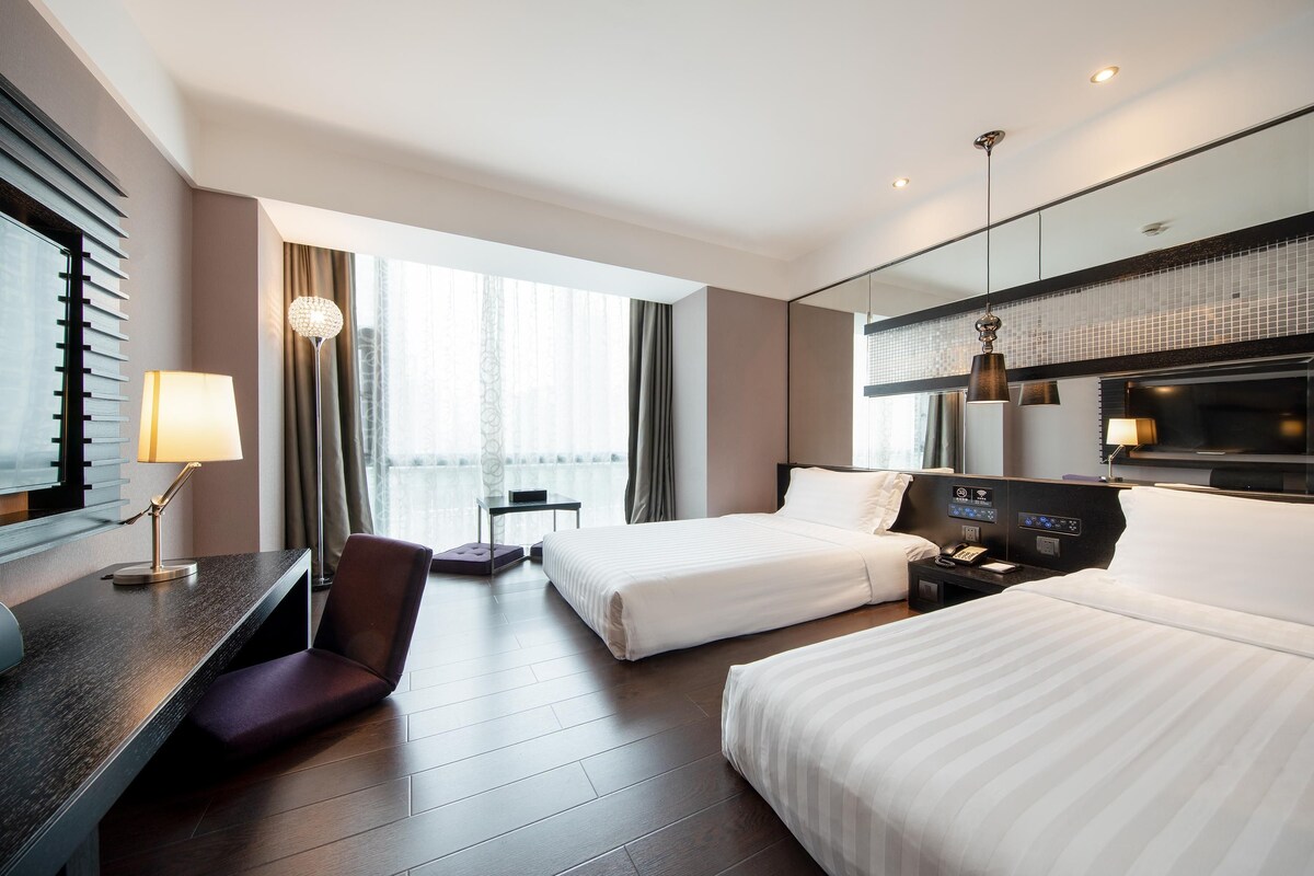Une chambre d’hôtel avec deux lits, un bureau avec une lampe, une chaise, un téléviseur et une grande fenêtre avec des rideaux.
