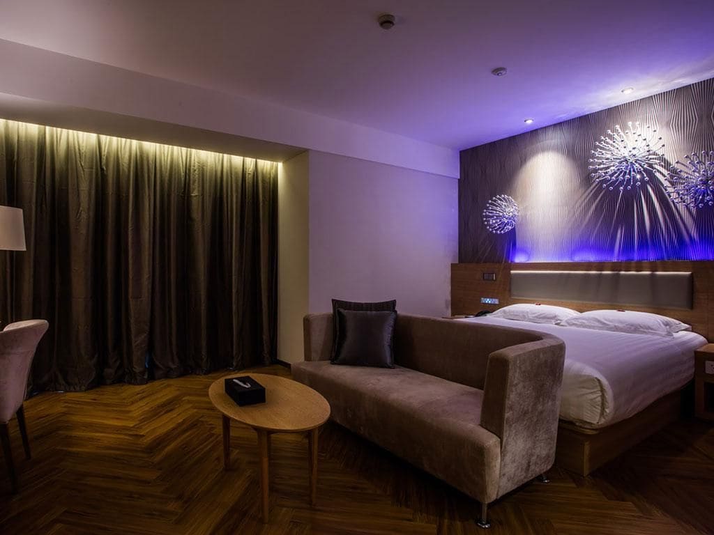 Una habitación de hotel moderna con una cama grande, un sofá, una mesita y una lámpara fijada a la pared.